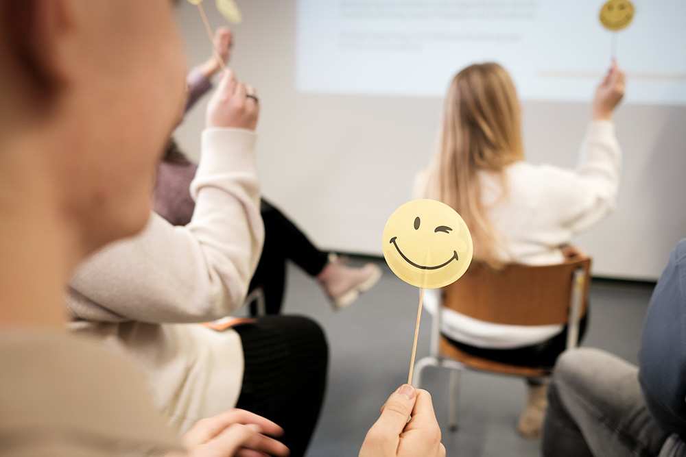Mehrere Personen sitzen in Richtung Leinwand in einem Klassenzimmer und halten Stäbchen mit lächelnden Gesichtern. (Quelle: Kindernothilfe)