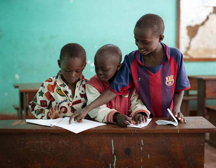 Drei Jungen an einem Schulpult, einer zeigt einem anderen etwas, indem er aufsteht und auf dessen Heft zeigt  (Quelle: Lars Heidrich)
