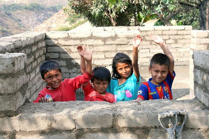 Kinder in Peru hinter einer weißen Mauer winken, im Hintergrund sieht man mehr von der weißen Mauer vor Bäumen und bergiger Gegend.  (Quelle: Jürgen Schübelin)